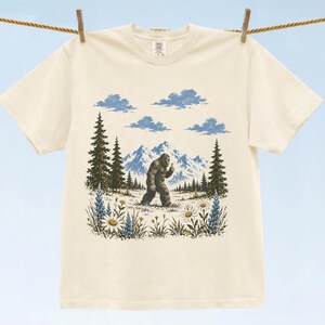 May include: A cream-colored short-sleeve t-shirt featuring a graphic print of a brown Bigfoot walking through a field of wildflowers with pine trees, blue mountains, and clouds in the background. The design is centered on the front of the shirt.