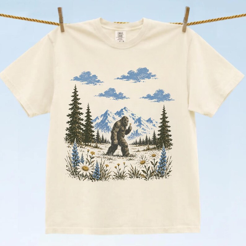 May include: A cream-colored short-sleeve t-shirt featuring a graphic print of a brown Bigfoot walking through a field of wildflowers with pine trees, blue mountains, and clouds in the background. The design is centered on the front of the shirt.
