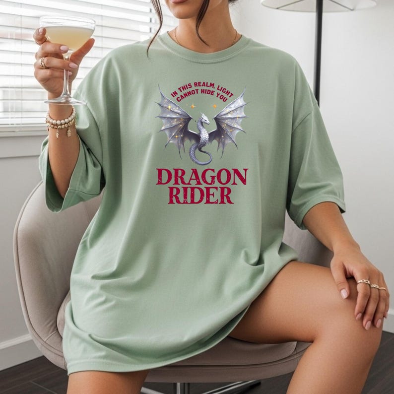 Puede incluir: Camiseta verde salvia con un gr&aacute;fico de drag&oacute;n. El gr&aacute;fico presenta un drag&oacute;n plateado con alas extendidas y el texto "IN THIS REALM LIGHT CANNOT HIDE YOU" encima de "DRAGON RIDER" en borgo&ntilde;a.