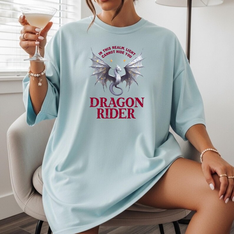 Puede incluir: Camiseta azul claro con un dise&ntilde;o de drag&oacute;n y el texto "IN THIS REALM, LIGHT CANNOT HIDE YOU" y "DRAGON RIDER". El drag&oacute;n es plateado con detalles morados y dorados. La camiseta es holgada.