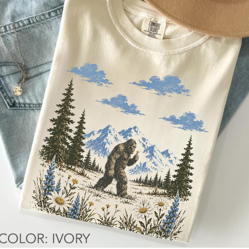 Bigfoot wildflower graphic t-shirt featuring a legendary Sasquatch walking through colorful mountain wildflowers, evergreen trees, and scenic alpine peaks in a vintage outdoor design on a unisex tee.