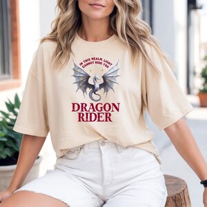 Puede incluir: Camiseta beige con un dise&ntilde;o de drag&oacute;n y el texto "IN THIS REALM, LIGHT CANNOT HIDE YOU" encima del drag&oacute;n y "DRAGON RIDER" debajo. El drag&oacute;n tiene alas plateadas y un dise&ntilde;o detallado.