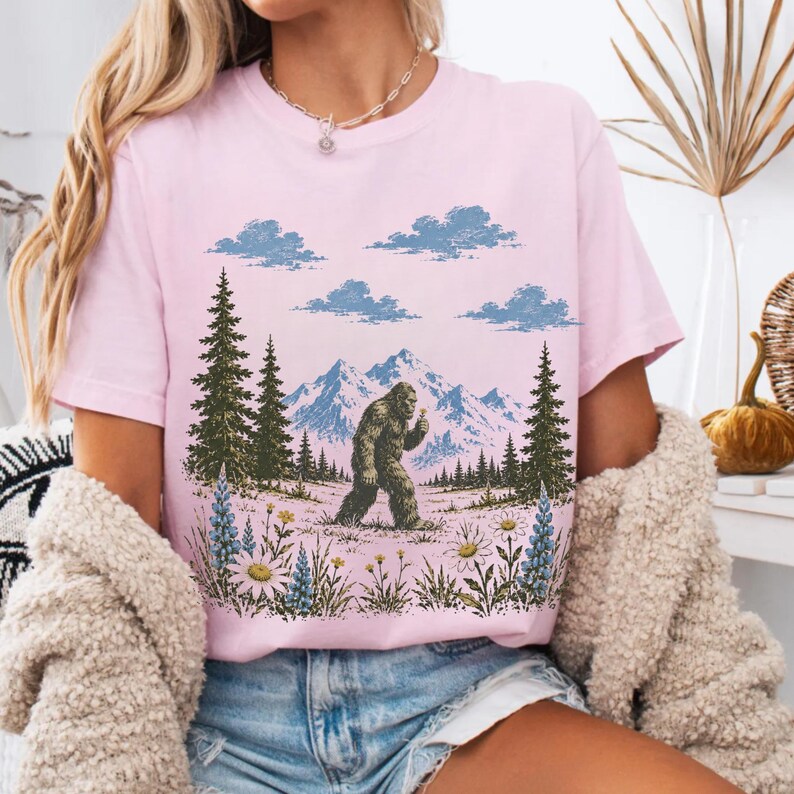 May include: A light pink short-sleeve t-shirt featuring a graphic print of a Bigfoot creature walking through a landscape with green pine trees, blue mountains, white clouds, and colorful wildflowers in the foreground.