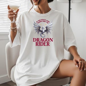Puede incluir: Camiseta blanca con un gr&aacute;fico de drag&oacute;n y el texto "IN THIS REALM LIGHT CANNOT HIDE YOU" arriba y "DRAGON RIDER" abajo. El drag&oacute;n tiene alas plateadas y un cuerpo gris. El texto es de color burdeos.