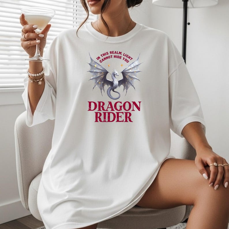 Puede incluir: Camiseta blanca con un gr&aacute;fico de drag&oacute;n y el texto "IN THIS REALM LIGHT CANNOT HIDE YOU" arriba y "DRAGON RIDER" abajo. El drag&oacute;n tiene alas plateadas y un cuerpo gris. El texto es de color burdeos.