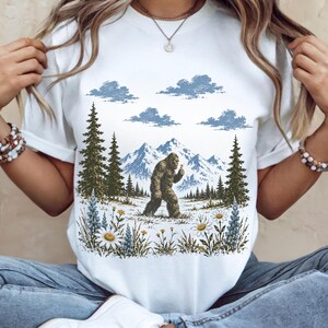 May include: A white t-shirt featuring a graphic print of a brown Bigfoot walking through a mountain landscape with pine trees, blue clouds, and a foreground of white daisies and blue lupine flowers. The design is centered on the chest area.