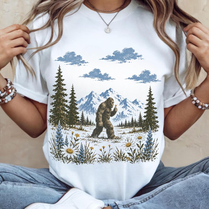 May include: A white t-shirt featuring a graphic print of a brown Bigfoot walking through a mountain landscape with pine trees, blue clouds, and a foreground of white daisies and blue lupine flowers. The design is centered on the chest area.