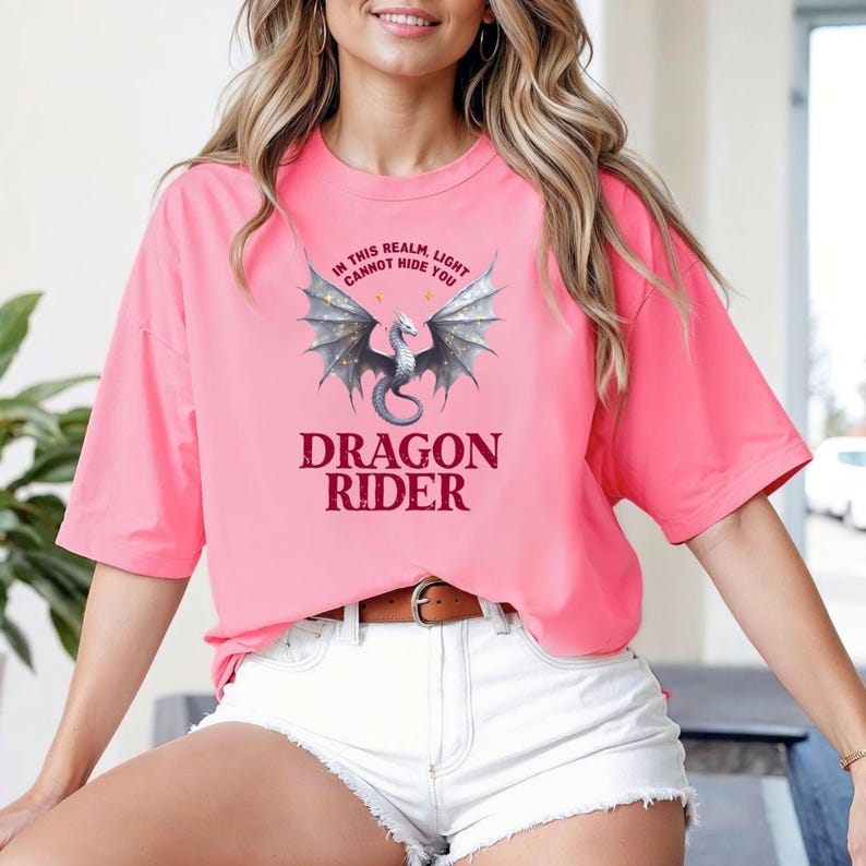 Puede incluir: Una camiseta rosa con un dise&ntilde;o de drag&oacute;n y el texto "IN THIS REALM, LIGHT CANNOT HIDE YOU" encima del drag&oacute;n y "DRAGON RIDER" debajo. El drag&oacute;n tiene alas grises y un cuerpo plateado.