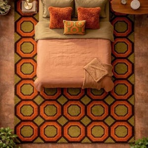 May include: A bedroom scene featuring a large area rug with a repeating orange and olive green geometric octagon pattern. The bed is dressed in a solid peach-coloured duvet, olive green pillows, two textured orange cushions, and a small patterned pillow.