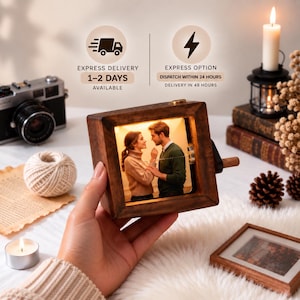 May include: A square wooden music box featuring a backlit photograph of a couple standing close together. The box has a small hand crank on the side and a warm internal light that illuminates the image of the two people within the frame.