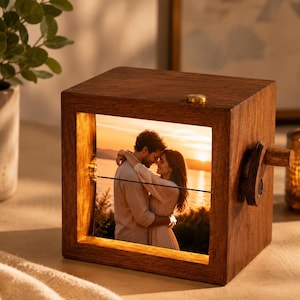 May include: A square wooden frame with a dark brown finish containing a backlit photograph of a couple embracing at sunset. The frame features a small brass-coloured knob on the top edge and a wooden crank handle on the right side for manual rotation.