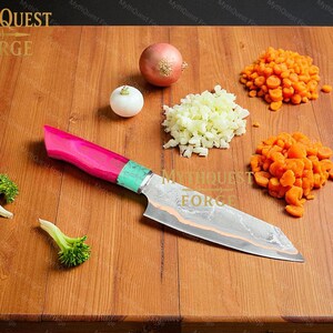 Pode incluir: Uma faca de chef com cabo rosa e verde repousa sobre uma t&aacute;bua de cortar de madeira. Cebolas picadas, cenouras e floretes de br&oacute;colis s&atilde;o dispostos ao redor da faca. A l&acirc;mina da faca tem um padr&atilde;o &uacute;nico. O logotipo MythQuest Forge &eacute; vis&iacute;vel.