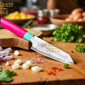 Pode incluir: Uma faca de chef de a&ccedil;o Damasco com uma pega rosa vibrante e turquesa repousa sobre uma t&aacute;bua de corte de madeira. A l&acirc;mina apresenta um padr&atilde;o intrincado. A cena inclui vegetais picados e o texto "MythQuest Forge".