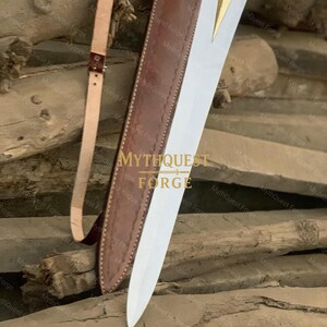 May include: A sword with a polished steel blade and a gold-colored hilt is partially sheathed in a brown leather scabbard. A tan leather strap is attached. The text "MYTHQUEST FORGE" is visible.