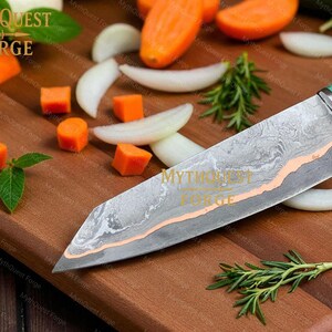Pode incluir: Uma faca de chef de a&ccedil;o Damasco com cabo verde e rosa repousa sobre uma t&aacute;bua de corte de madeira. A l&acirc;mina tem um padr&atilde;o &uacute;nico com um detalhe cor de cobre. Cenouras picadas, rodelas de cebola e alecrim circundam a faca. O texto "MYTHQUEST FORGE" &eacute; vis&iacute;vel.