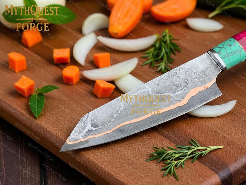 Pode incluir: Uma faca de chef de a&ccedil;o Damasco com cabo verde e rosa repousa sobre uma t&aacute;bua de corte de madeira. A l&acirc;mina tem um padr&atilde;o &uacute;nico com um detalhe cor de cobre. Cenouras picadas, rodelas de cebola e alecrim circundam a faca. O texto "MYTHQUEST FORGE" &eacute; vis&iacute;vel.
