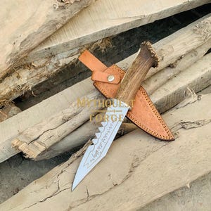 Könnte beinhalten: Ein handgefertigtes Messer mit gezahnter Klinge und Hirschhorngriff liegt in einer braunen Lederscheide. Die Klinge hat geätzten Text. Das Messer und die Scheide liegen auf einer Holzoberfläche. Der Text "MythQuest Forge" ist sichtbar.