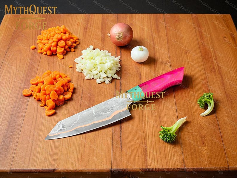 Pode incluir: Uma faca de chef com cabo rosa e verde repousa sobre uma t&aacute;bua de cortar de madeira, junto com cenouras picadas, cebolas e floretes de br&oacute;colis. A l&acirc;mina da faca tem um padr&atilde;o &uacute;nico. O texto "MythQuest Forge" &eacute; vis&iacute;vel.