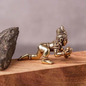 Könnte beinhalten: Kleine, goldfarbene Figur einer knienden Gottheit, die eine Kugel hält. Die Figur steht auf einem rechteckigen Holzblock, mit einem dunkelgrauen Stein links. Der Hintergrund ist ein gedämpftes Grau.