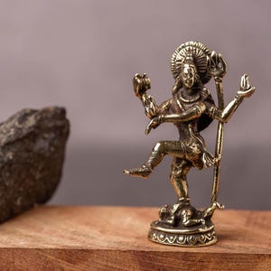 Puede incluir: Una pequeña estatua dorada de una deidad hindú, posiblemente Shiva, en pose de baile. La estatua está sobre un bloque de madera con una piedra rugosa a la izquierda. El fondo es gris apagado.