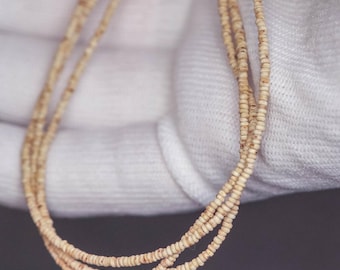 May include: A delicate, multi-strand necklace with small, light-colored beads. The necklace is held by a gloved hand against a dark background. The beads appear to be uniform in size and shape, creating a simple yet elegant design.
