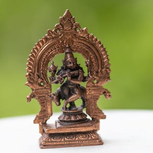 May include: A bronze-colored statue of a deity playing a flute, set within an ornate arch. The statue is on a white marble-topped stand with a wooden base, against a blurred green background. The statue is approximately 4 inches tall.