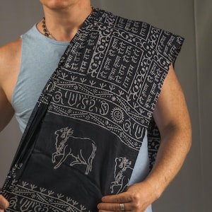 May include: A black and white patterned shawl featuring script and cow designs. The shawl is draped over the shoulder of a person wearing a light blue vest and white trousers.