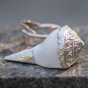 Puede incluir: Una concha blanca con detalles metálicos plateados y un anillo a juego. La concha tiene intrincados patrones en la parte superior y una punta plateada texturizada. Los artículos se exhiben sobre una superficie de piedra gris.