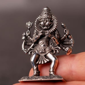 May include: A detailed silver figurine of a multi-armed deity, possibly Hindu, with a fierce expression and ornate headdress. The figure is standing on a small rectangular base. The figurine is held in a person's hand.