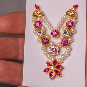 Könnte beinhalten: Eine goldfarbene Halskette mit Kunstperlen, rosa und klaren Edelsteinen und einem floralen Anhänger. Die Halskette wird auf einer weißen Karte präsentiert und in einer Hand gehalten.