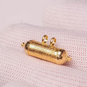 May include: A cylindrical gold pendant featuring a textured, cross-hatched surface pattern. The pendant has rounded ends with small protruding spheres and two small, vertical loops attached to the top center for hanging on a chain or cord.
