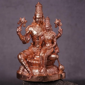 May include: A copper-coloured statue of two figures, one seated and the other standing, both with elaborate headdresses and holding objects. The statue is set against a dark grey background.