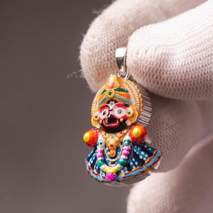 Puede incluir: Un colgante colorido con una figura de deidad con un diseño detallado. El colgante tiene una fianza plateada y está adornado con colores vibrantes, incluyendo rojo, azul, amarillo y verde. La figura lleva una corona y un vestido estampado.