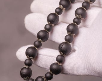 Puede incluir: Un collar oscuro de cuentas con cuentas grandes y redondas. Las cuentas son de color negro intenso y están ensartadas con detalles metálicos decorativos. El collar se sostiene en una mano enguantada sobre un fondo neutro.