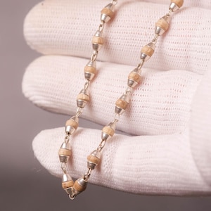 May include: A silver necklace featuring light brown, barrel-shaped beads linked together with silver. The necklace is held in a gloved hand against a neutral background. The beads are approximately 0.5 cm in diameter.