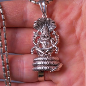 May include: A silver pendant necklace featuring a detailed deity figure seated atop a coiled serpent. The pendant has intricate detailing and is suspended from a silver chain. The design includes a cobra hood and multiple arms.