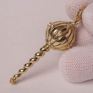 May include: A gold-toned pendant shaped like a mace, featuring a spherical head with vertical ridges and small embedded clear stones. The handle has a twisted, spiral design, and a small loop at the top allows for attachment to a chain or cord.