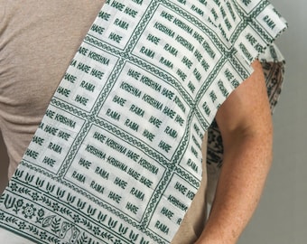 May include: White and green scarf with repeating text that reads "Hare Krishna Hare Rama". The scarf has a patterned border and is draped over the shoulder. The scarf is made of a lightweight fabric.