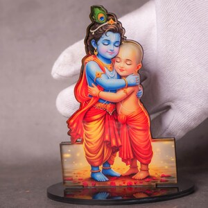 Peut inclure: Figurine en bois colorée représentant deux personnages enlacés. L'un est bleu avec une plume de paon dans les cheveux, l'autre est orange. La figurine est sur une base noire.