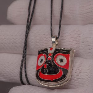 Op de afbeelding: Zilveren hangerketting met een rechthoekige hanger. De hanger heeft een zwart, rood en wit gezichtsontwerp. De ketting is donkergrijs. Een opvallend sieraad.