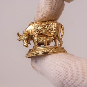 Könnte beinhalten: Eine kleine goldfarbene Metallfigur, die eine Kuh und ein Kalb auf einem runden, strukturierten Sockel darstellt. Die Kuh weist auf ihrem Rücken komplizierte florale Prägemuster auf, während der Sockel einen dekorativen Rand am Umfang hat.