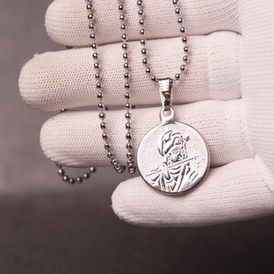 Peut inclure: Collier pendentif argenté avec un pendentif rond représentant une personne jouant de la flûte. Le collier est composé d'une chaîne à boules et est tenu par une main gantée.