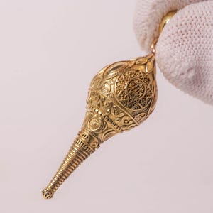 May include: A gold-colored pendant shaped like a conch shell, featuring intricate embossed patterns and a tapered, ribbed lower section. The surface is covered in detailed scrollwork and circular motifs, with a small loop at the top for attachment.