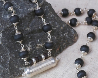 May include: A necklace with black, matte beads and silver-coloured metal detailing. The necklace includes a cylindrical pendant and is displayed on a textured grey stone. The beads are linked by fine silver chains, creating a stylish accessory.
