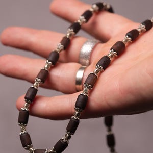 Puede incluir: Un collar de cuentas marrón oscuro con detalles plateados. El collar presenta cuentas cilíndricas ensartadas con conectores metálicos decorativos. La joya se sostiene en una mano sobre un fondo neutro.