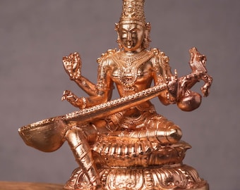 May include: A bronze-colored statue of a seated deity, adorned with intricate details and multiple arms. The figure holds a stringed instrument and is seated on a lotus flower base. The statue is placed on a wooden block.