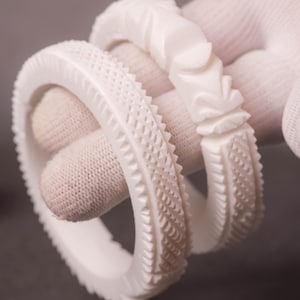 Könnte beinhalten: Drei weiße, aufwendig geschnitzte Armreifen. Die Armreifen weisen detaillierte Muster auf, darunter strukturierte Rippen und florale Designs. Der Schmuck wird von einer behandschuhten Hand gehalten, die die Handwerkskunst und das Design der Armbänder zeigt.