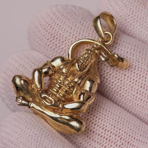May include: A gold-toned pendant featuring a detailed figure seated in a meditative pose with legs crossed, arms resting on the knees, and a curved element extending from the head, finished with a small circular bail at the top for attachment.