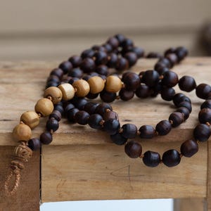 May include: Two wooden prayer bead necklaces rest on a small, rustic wooden stool. One necklace features dark brown beads, while the other has a mix of light and dark brown beads. The beads are strung on a cord.