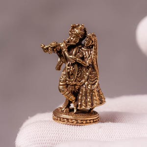 May include: A small, gold-colored statue depicting two figures, one playing a flute. The figures are adorned with detailed clothing and stand on a decorative base. The statue is held in a white glove.